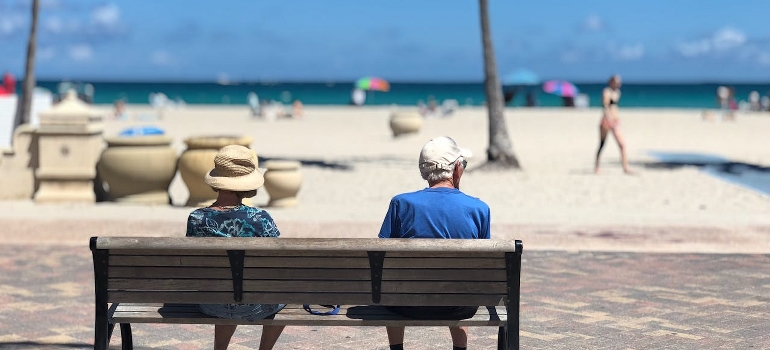 Two seniors are sitting on a brown wooden bench in Miami Beach.