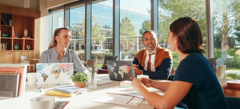 Three colleagues engaged in a business meeting, discussing plans in a bright, modern office setting with large windows.