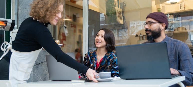 a man and a woman with laptop sitting outside of the coffee shop and getting coffee served