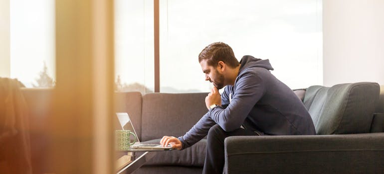 A man sitting on the couch and reading about how to approach moving to the USA from Canada on a laptop