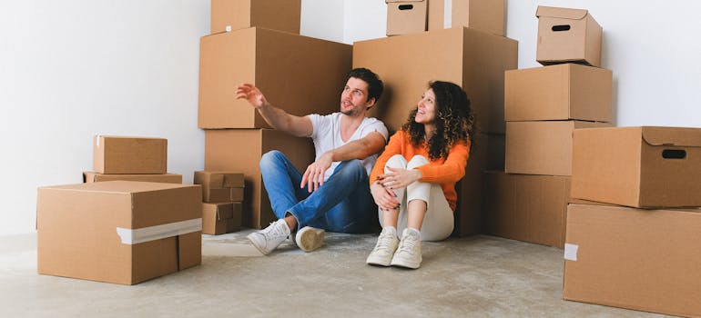 a man and a woman sitting on the floor among cardboard boxes and discussing