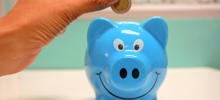 A close-up of a person dropping a coin into a smiling blue piggy bank, symbolizing cost-saving strategies for residential, long-distance, and interprovincial relocations in Canada.