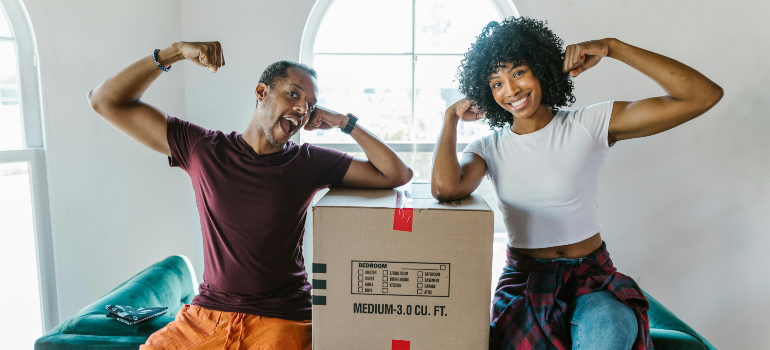 A cheerful couple flexing their arms while sitting next to a packed moving box, celebrating a successful move with residential, long-distance, and interprovincial relocations in Canada.