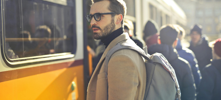 A stylish man with a backpack waiting near a bus, looking into the distance as people board the vehicle.
