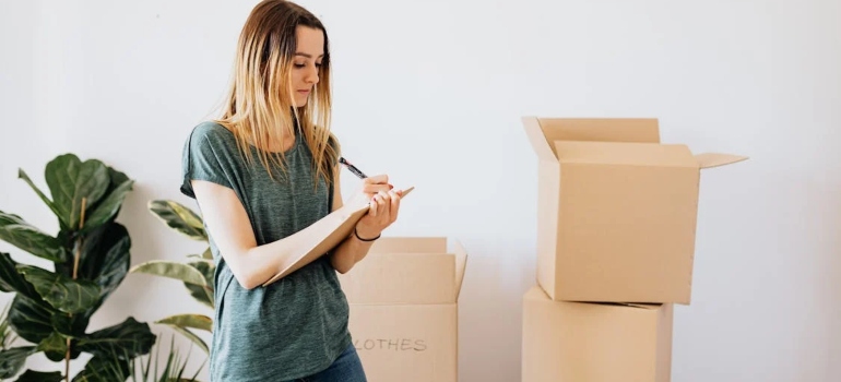 A woman writing on a clipboard estimating whether decluttering before a move feels so emotionally liberating, in the background there are boxes and plants