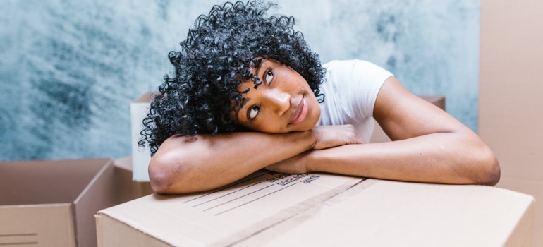 A woman with curly hair leaning on her head on a box thinking about how decluttering before a move feels so emotionally liberating