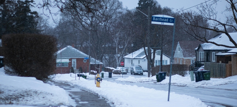 A snowy residential street lined with modest homes and garbage bins, ideal for highlighting the Dos and Don'ts of Your Long-Distance Move.