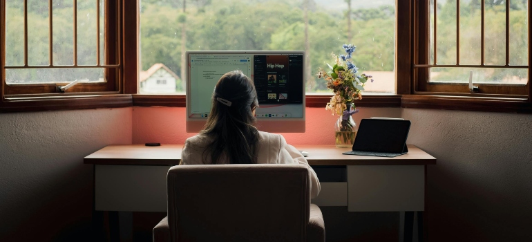 A woman working at a desktop computer with a peaceful view, symbolizing the question of How Remote Work Is Affecting Moving Trends in Ontario.