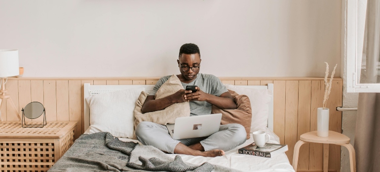 A man comfortably working from bed using his laptop and phone during remote work hours.
