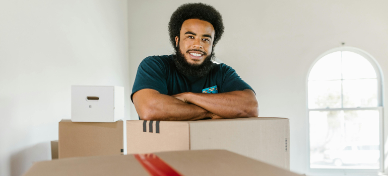 Smiling mover leaning on stacked boxes in a bright room, reflecting service quality and traits of reliable cross border moving companies.