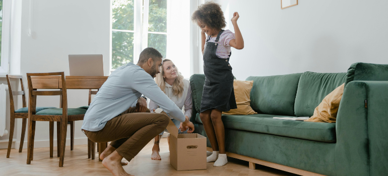 Parents kneel by a box while their daughter cheers in the living room, packing together to prepare your family for a cross border move.
