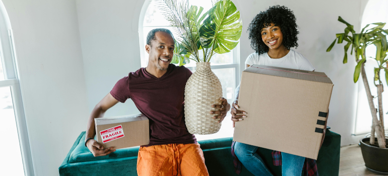 A smiling couple sits at home with boxes and a houseplant as they choose between hiring movers vs. DIY.