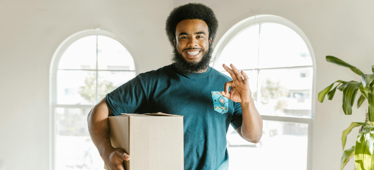 Mover holding a box and giving an OK sign in a bright room, trained crew and courteous service, characteristics of a reliable moving & packing company.