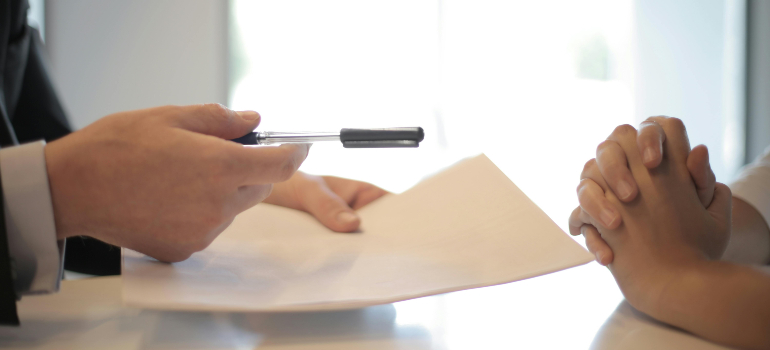 Hands at a desk during paperwork, with one person offering a pen over forms while another waits with clasped hands.