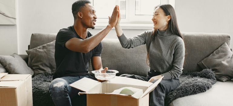 Couple on a sofa high-fiving while unpacking boxes, showing organization, care, and teamwork, characteristics of a reliable moving & packing company.