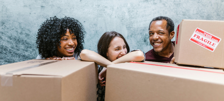 Three friends smiling behind boxes labeled FRAGILE, clear labels and gentle handling, characteristics of a reliable moving & packing company.