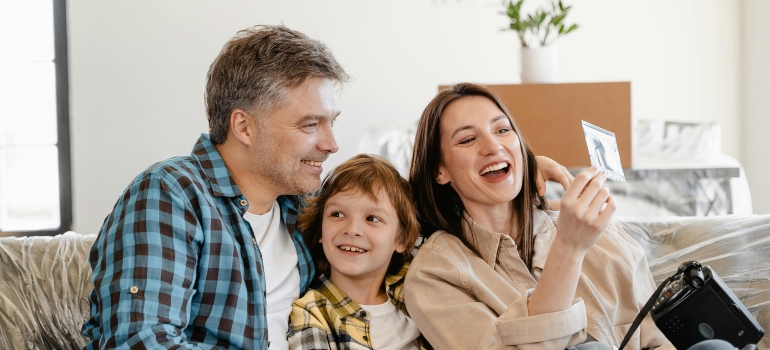 Family taking a photo together in their new home while unpacking boxes, with movers working safely in the background.