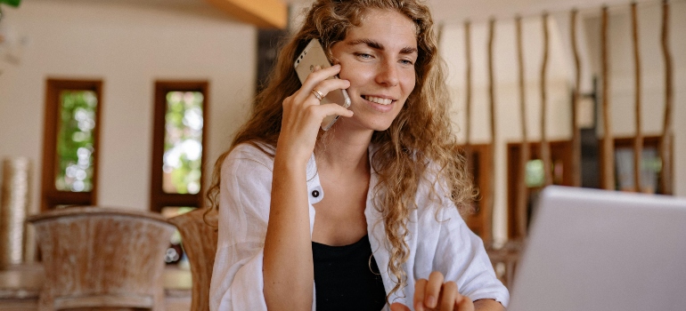 Smiling woman on the phone and using a laptop to call the moving company and find out what can you do when your movers are late.