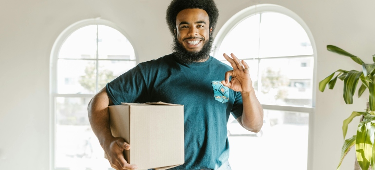 Professional mover smiling and holding a cardboard box, giving an OK hand sign in a bright room with large windows.