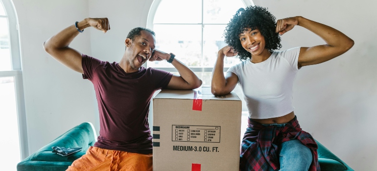 A couple smiling and flexing beside a packed moving box after delivery with movers Markham residents adore.
