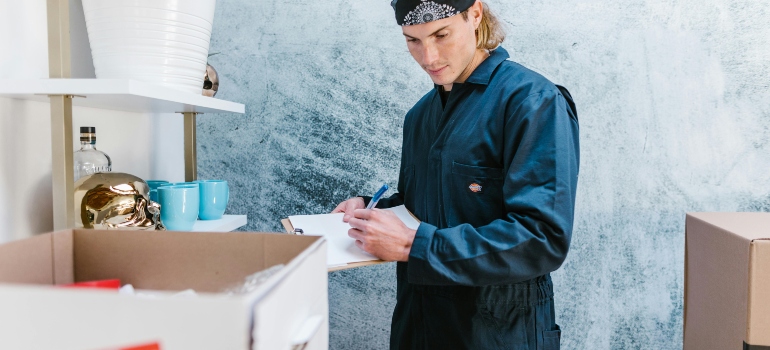 Mover writes a checklist beside packed boxes and shelving while preparing for a workplace relocation.