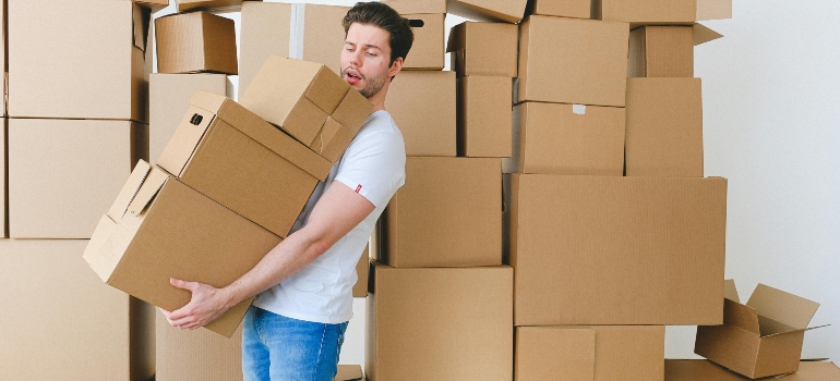 Man lifting multiple moving boxes in front of stacked cartons, demonstrating physical demands of relocation.